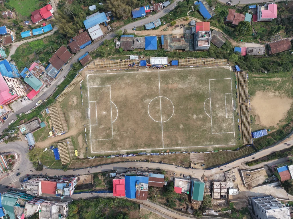 Aerial view of Ilam Tudikhel football ground in Ilam Bazaar, Nepal, surrounded by buildings, roads, and green hillsides under daylight.