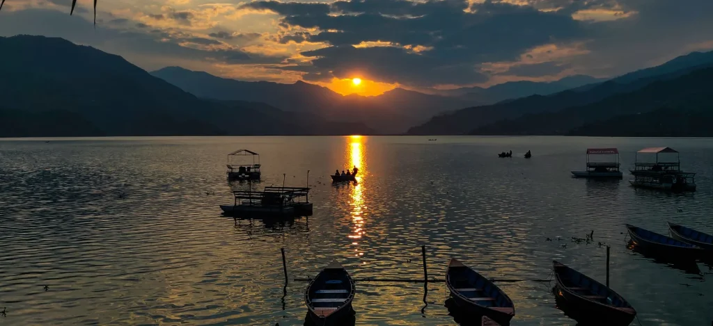 Golden sunset over Phewa Lake in Pokhara, Nepal, with silhouettes of traditional wooden boats in the foreground and mountains under a cloudy sky.