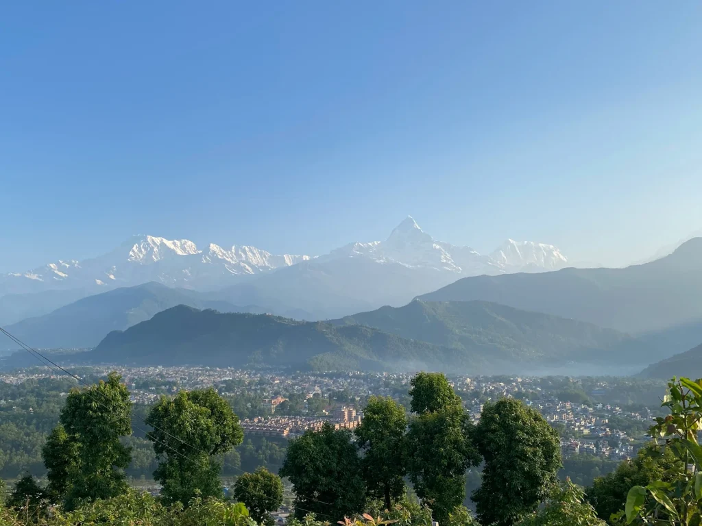 Panoramic view from Sarangkot viewpoint looking down at Pokhara city valley, with the towering snow-covered Annapurna mountain range and Machhapuchhre peak under a clear blue sky.