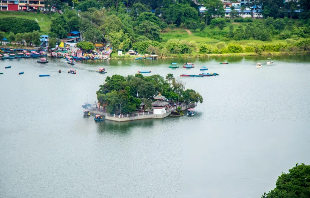 An elevated, high-angle aerial view looking down at Tal Barahi Temple island on the calm waters of Phewa Lake, showcasing the mainland Lakeside docks, scattered colorful rowboats, and the vibrant shoreline of Pokhara, Nepal.