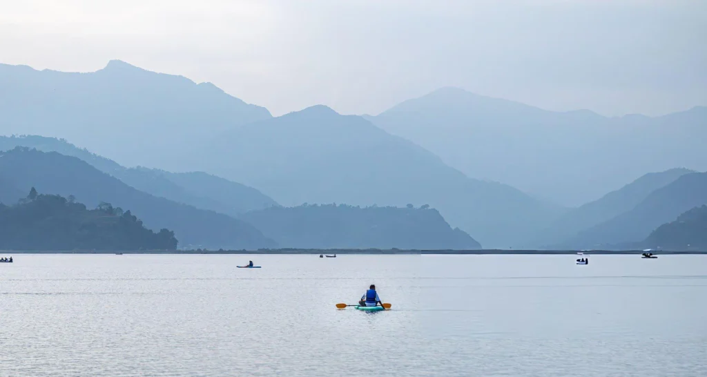 A peaceful, minimalist landscape photograph of a single adventurer kayaking on the vast, calm waters of Phewa Lake in Pokhara, Nepal, with layers of soft, misty blue mountain ridges fading into the background sky.