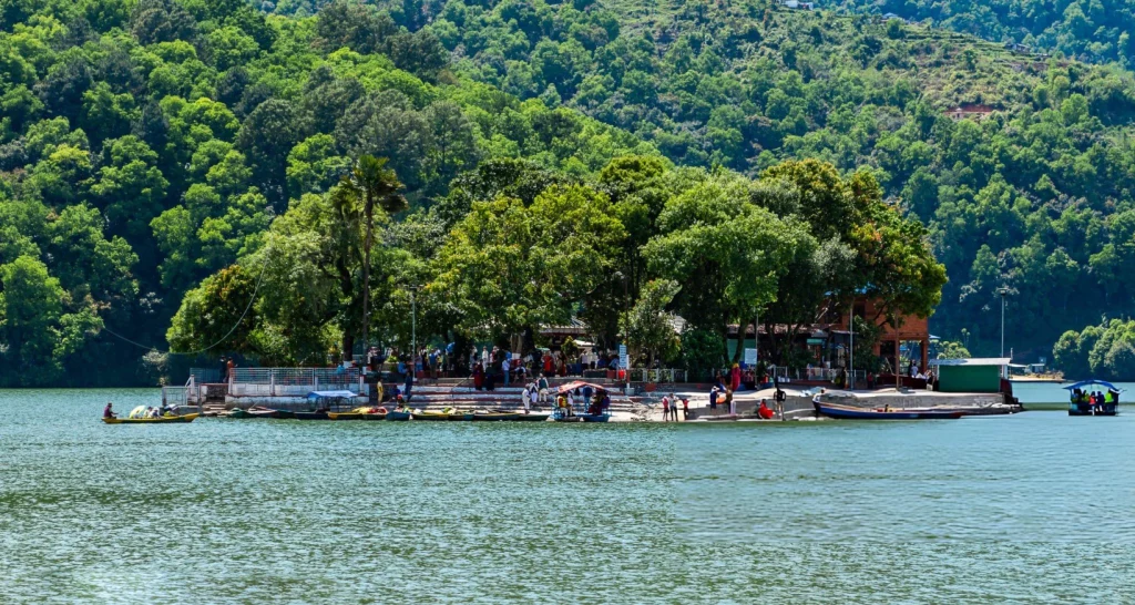 A vibrant daytime shot of the historic, multi-tiered Hindu shrine Tal Barahi Temple situated on a small, lush green island in the middle of Phewa Lake, surrounded by parked colorful wooden rowboats and a forested hill backdrop in Pokhara, Nepal.